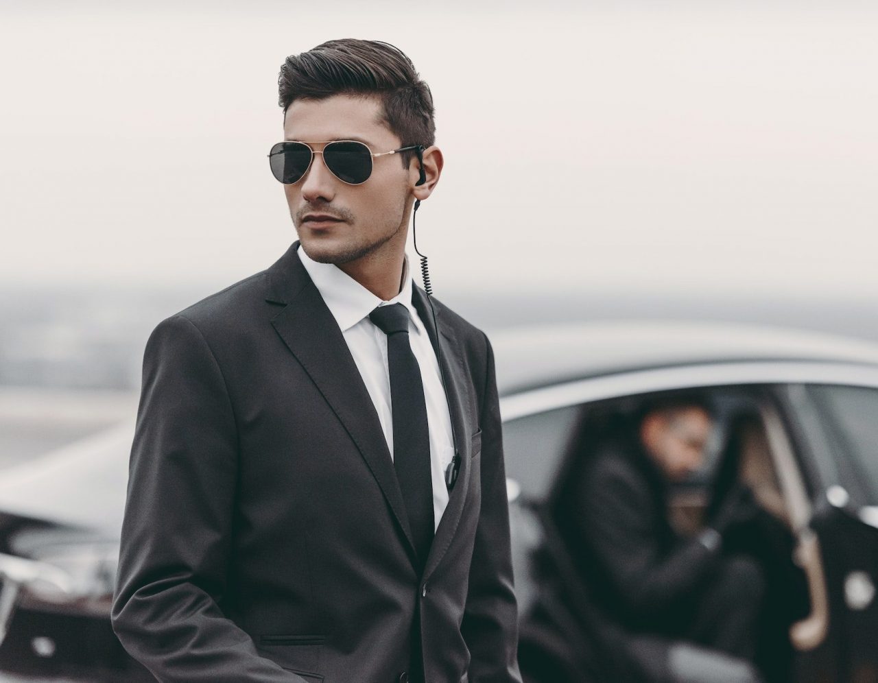 A man in a black suit, tie, and sunglasses with an earpiece stands alert in front of a black car, representing top-tier Security Services in Oakland County, Michigan and another person is stepping out of the car in the background.
