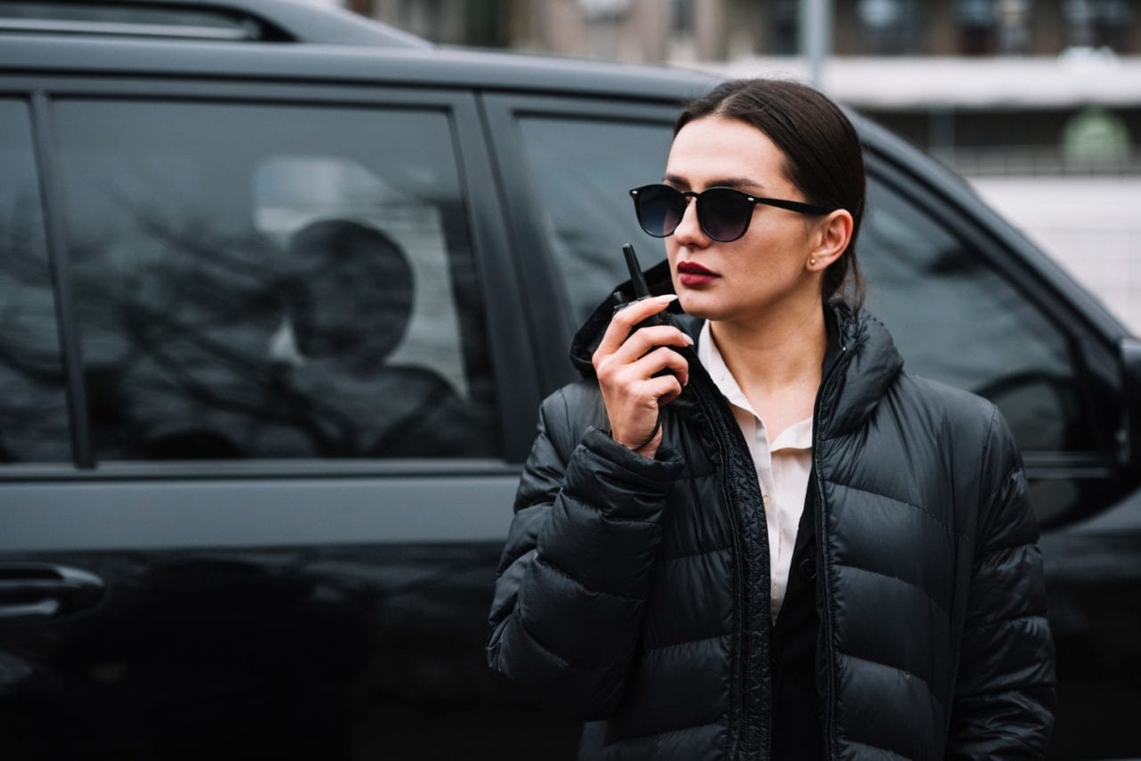 A woman wearing sunglasses and a black puffer jacket stands next to a black vehicle, holding a walkie-talkie near her mouth, appearing alert and focused while providing security services in Oakland County, Michigan.