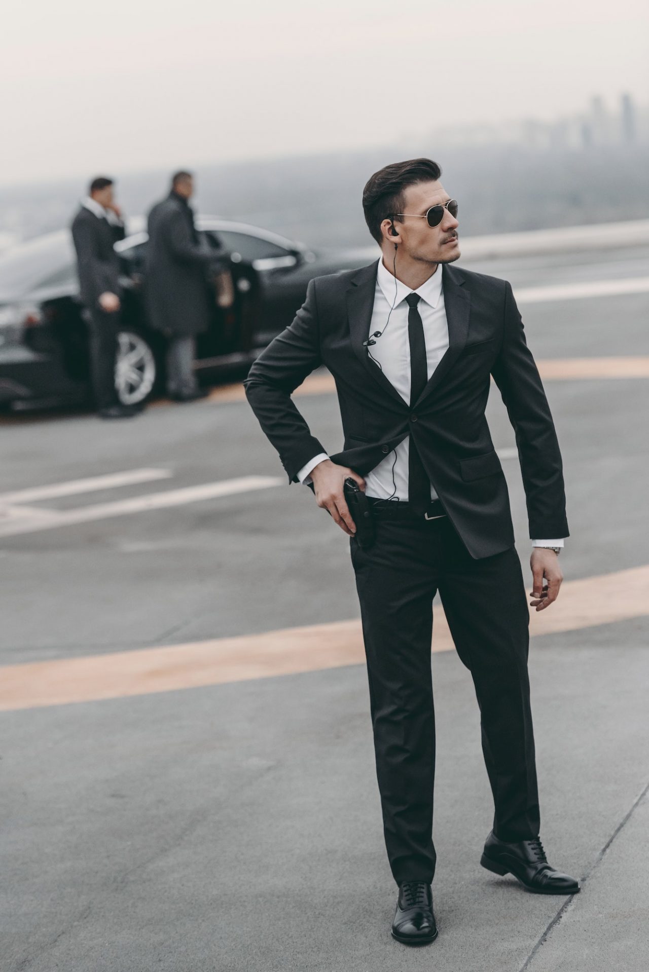 A man in a black suit and sunglasses, wearing an earpiece, stands alert on a rooftop near a black car. Two other suited men talk by the car in the background, highlighting professional Security Services on duty.