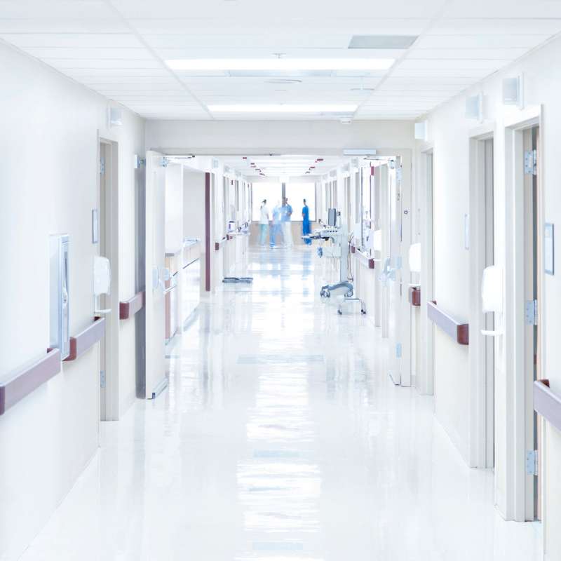 A bright, sterile hospital corridor with white walls and shiny floors stretches ahead. Several doors line the hallway, while a few medical staff in scrubs engage in quiet conversation. Nearby, Private Security Oakland County ensures safety and peace within the bustling environment.
