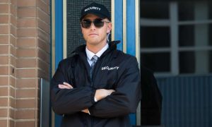 A security guard wearing a black jacket, cap, and sunglasses stands with crossed arms. The word "SECURITY" is emblazoned on his attire. Positioned in front of a brick wall and a window, he exemplifies the vigilant presence of Security Services Oakland County.