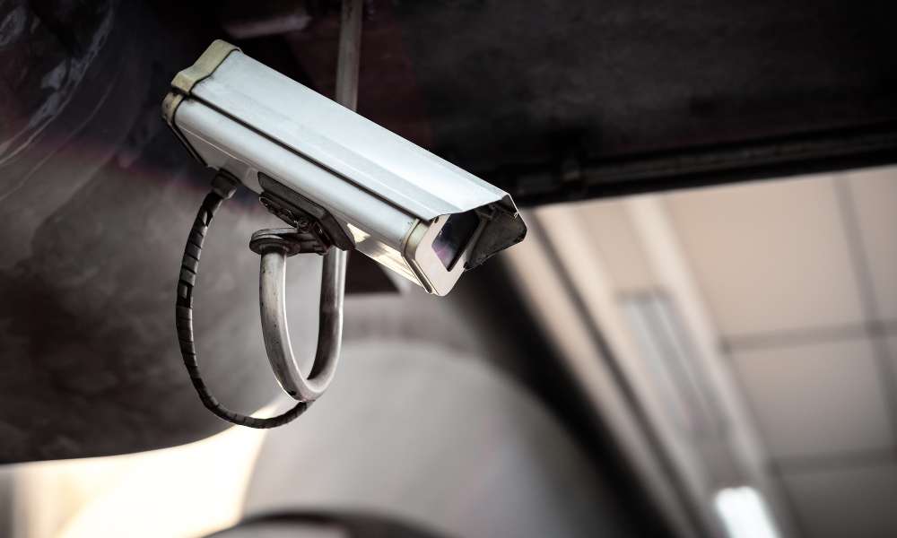 A white security camera, installed by a leading Security Company in Oakland County, rests on a metal bracket under the ceiling. Angled downward, with black wiring, it surveys the dimly lit indoor environment where background elements blur into obscurity.