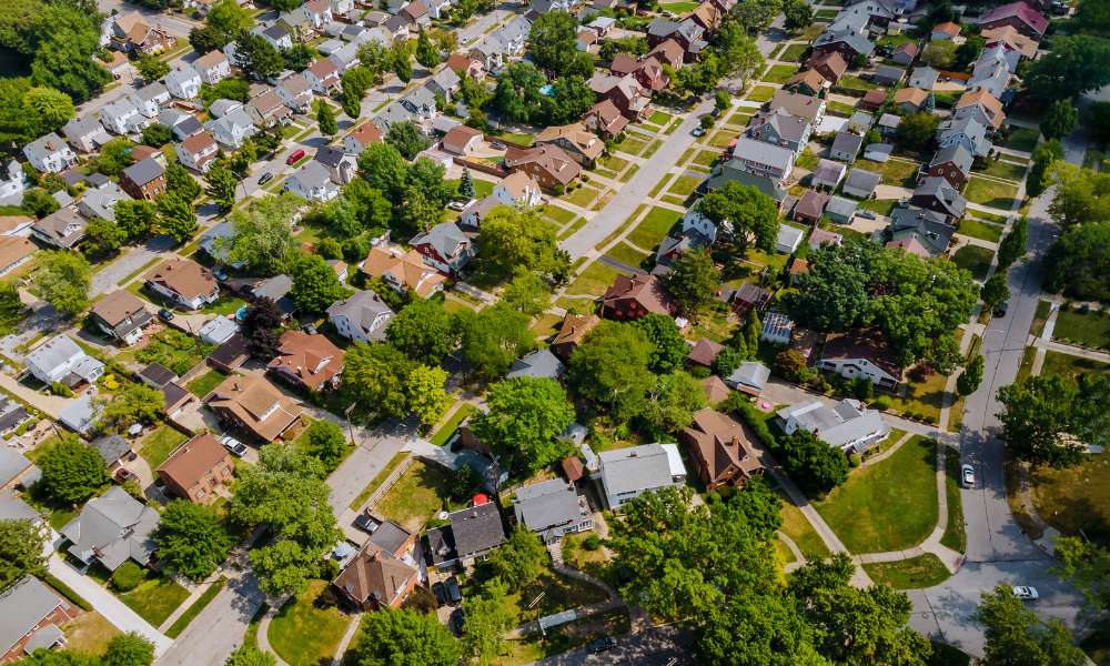 Aerial view of a suburban neighborhood in Oakland County, featuring neatly arranged houses, tree-lined streets, and lush greenery. The area reflects a sense of community with various home styles and well-maintained lawns, where security companies ensure peace during special events.