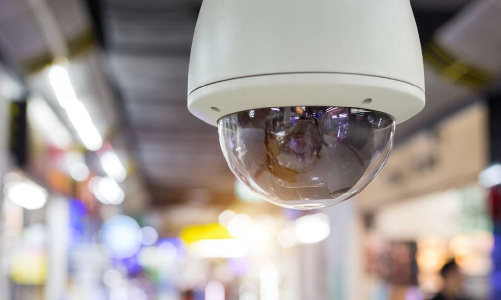 A close-up of a dome-shaped security camera in a brightly lit indoor setting, possibly a shopping mall or public space in Oakland County. The background is blurred, highlighting colorful lights and signs, perfectly capturing the vigilance provided by Private Security Oakland County.