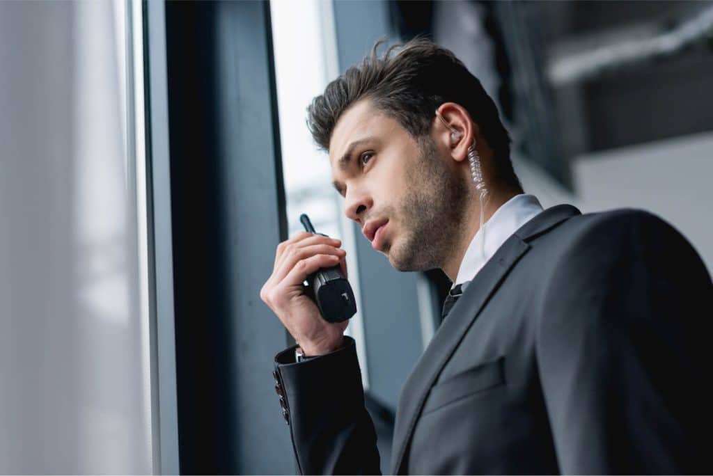 A man in a suit with an earpiece and walkie-talkie stands alert by a window, providing Security Services in Oakland County, Michigan.