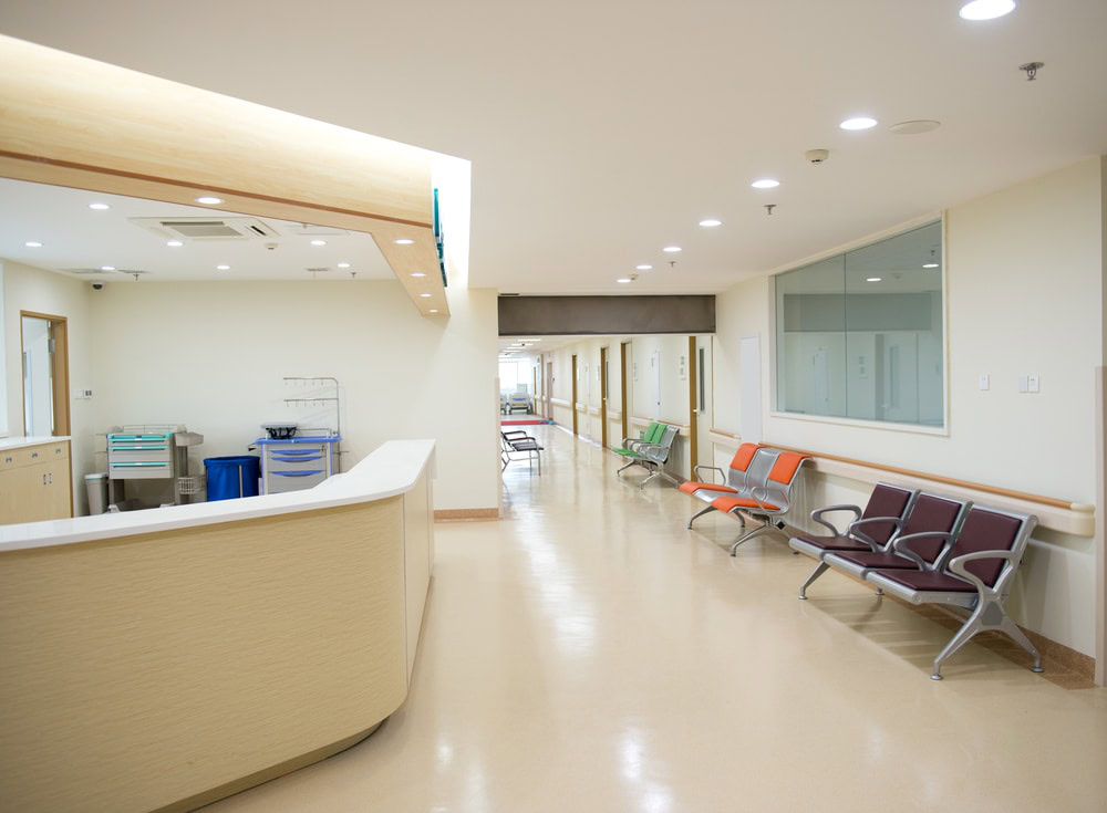 A hospital corridor with beige flooring and walls features a reception counter on the left. Several rows of colorful chairs are placed along the walls, while bright overhead lights illuminate the area, creating a clean and orderly environment. Private Security Oakland County ensures safety throughout the space.