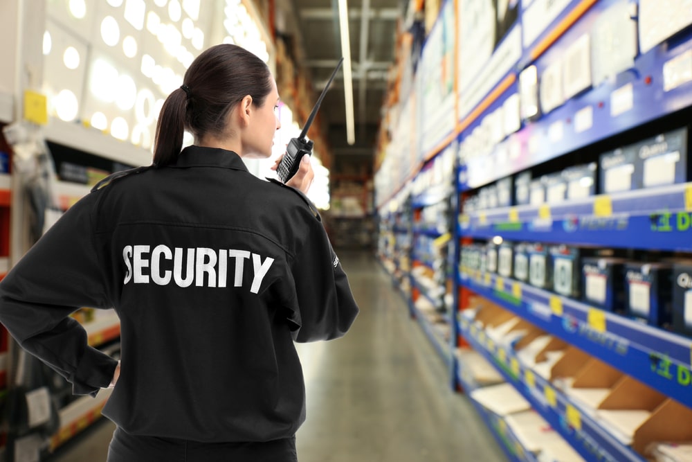 A security guard wearing a black uniform with "SECURITY" on the back and representing a top private security company in Oakland County holds a walkie-talkie, standing in the aisle of a large warehouse. Shelves stocked with various products are visible on the right side.
