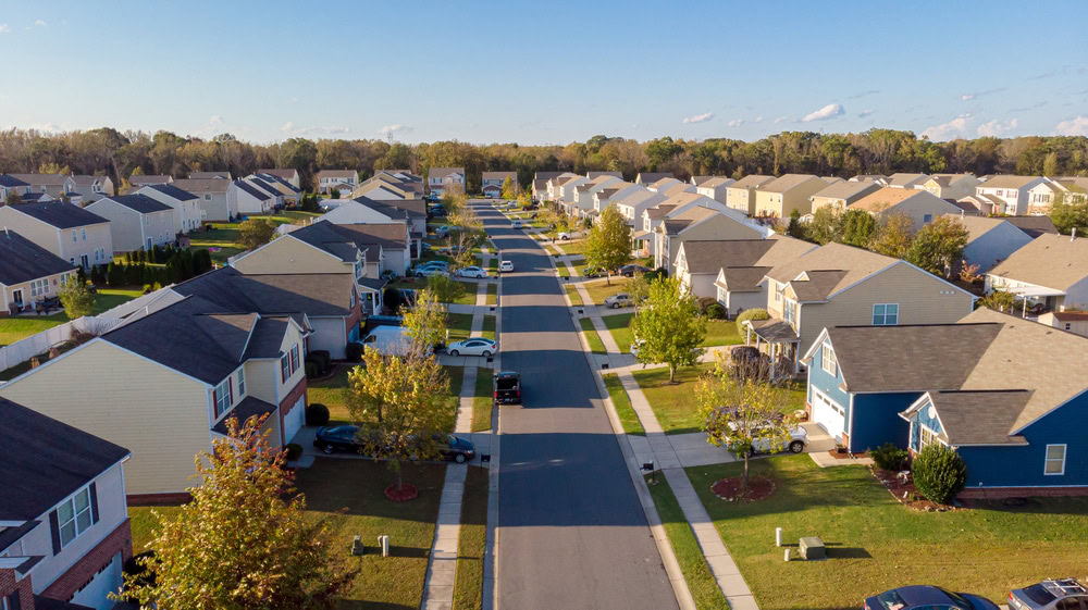 Aerial view of a suburban neighborhood in Oakland County, where rows of similar single-family homes feature sloped roofs and small front yards with neatly trimmed grass. A long, straight road runs between the houses, lined with trees, parked cars, and the assurance of private security from a local company.