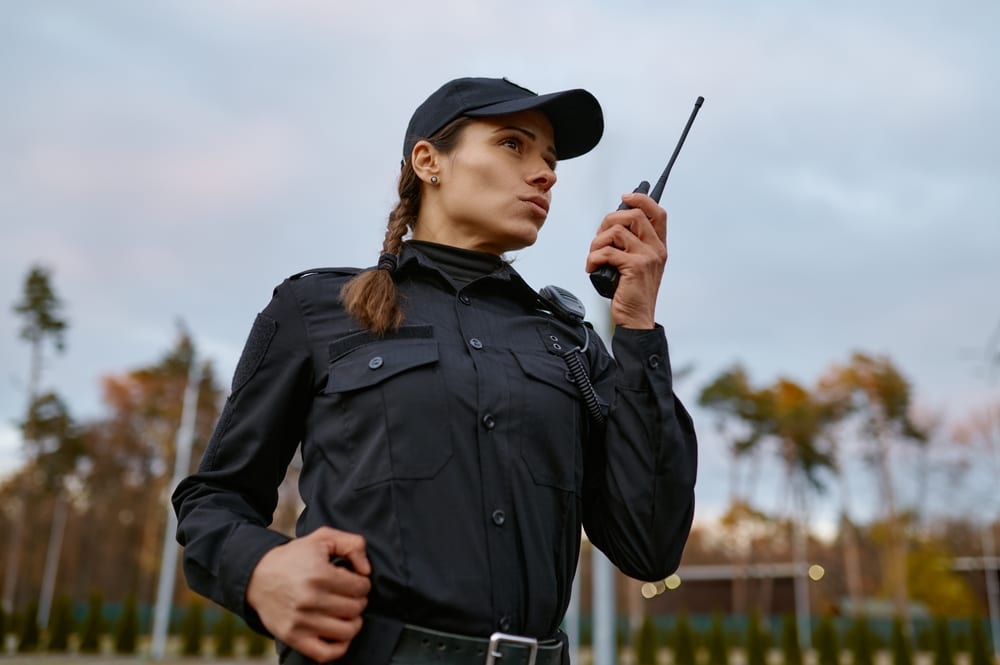 A security officer in uniform stands outdoors, holding a walkie-talkie to her mouth. She is focused on communicating, representing a Security Company in Oakland County, with trees and a cloudy sky in the background.