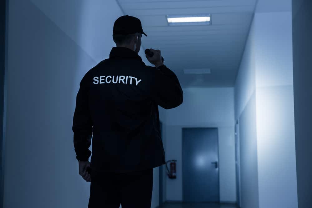 A security guard in uniform holds a flashlight while standing in a dimly lit hallway, proudly representing Private Security Oakland County. The word "SECURITY" is visible on the jacket's back. The narrow hallway has doors on either side and a light fixture above, ensuring safety during special events.