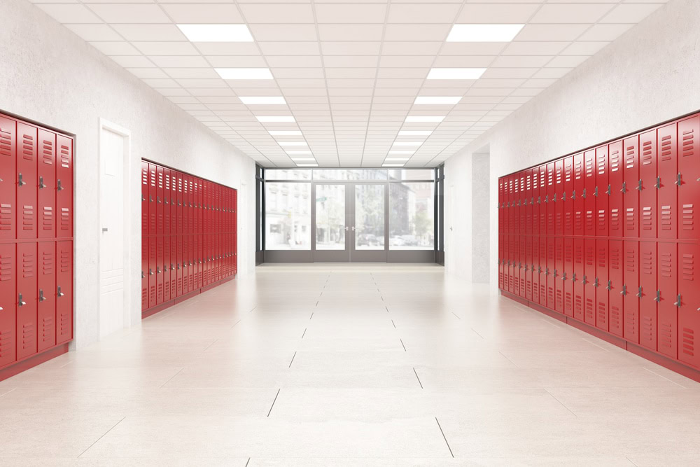 A long hallway with red lockers lining both sides leads to large windows at the end. The ceiling's white tiles and the light beige floor create a bright, spacious school corridor, monitored discreetly by private security for Oakland County's safety standards.