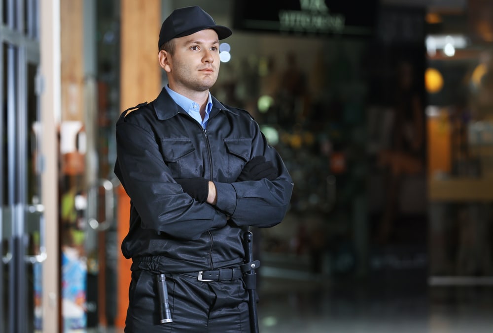 A security guard in a dark uniform and cap stands with arms crossed, vigilantly overseeing the bustling shopping mall. The backdrop is slightly blurred, highlighting various shops and lights. This scene exemplifies the professionalism of private security in Oakland County during special events.