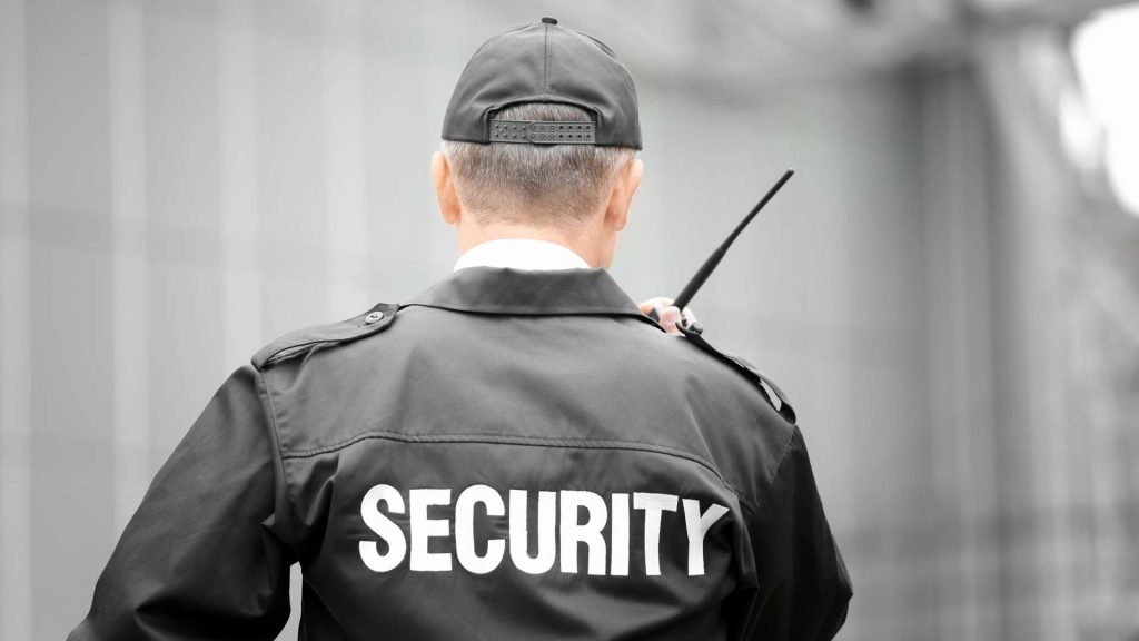 A security guard in a black uniform and baseball cap labeled "SECURITY" holds a walkie-talkie. Facing away from the blurred, modern building facade, he represents Private Security in Oakland County with vigilance and professionalism.
