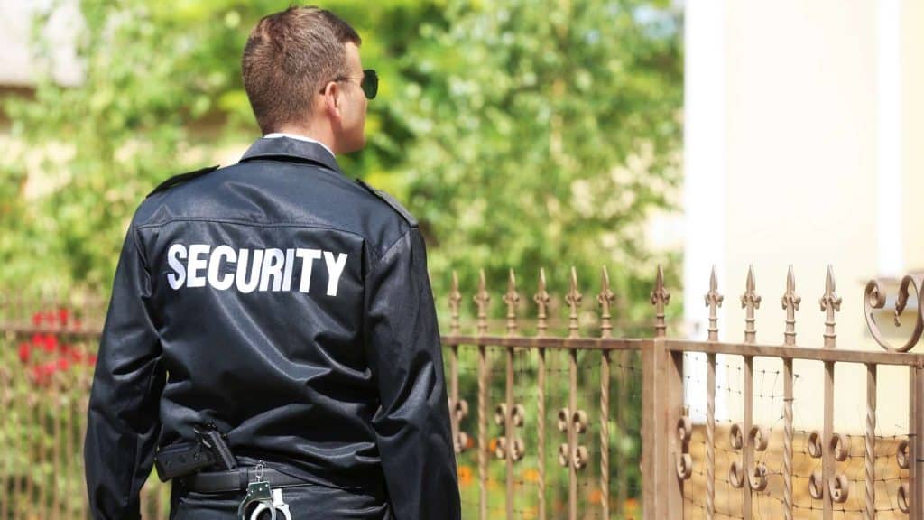 A security guard wearing a black uniform and sunglasses stands outdoors, with "SECURITY" emblazoned on the back of his jacket. He surveys a wrought iron fence, greenery in the background, representing top-tier Private Security Oakland County ensures for any environment.