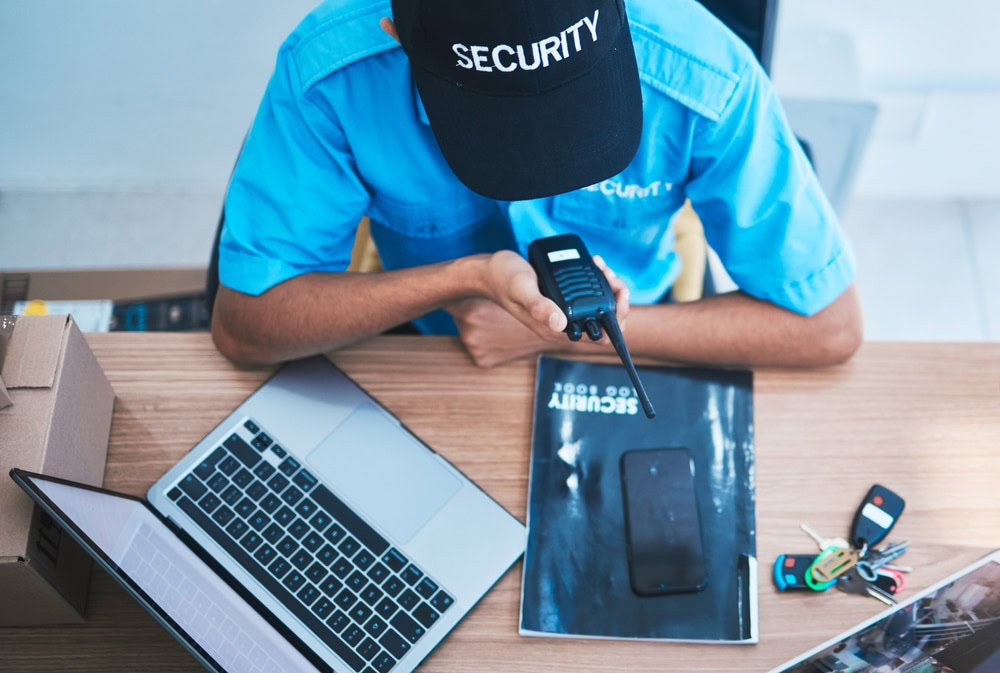 A security guard, part of a leading Security Company in Oakland County, sits at a desk with a laptop, two-way radio, keys, and cell phone. Dressed in a blue uniform and black "SECURITY" cap, the guard speaks into the radio while vigilantly overseeing operations.
