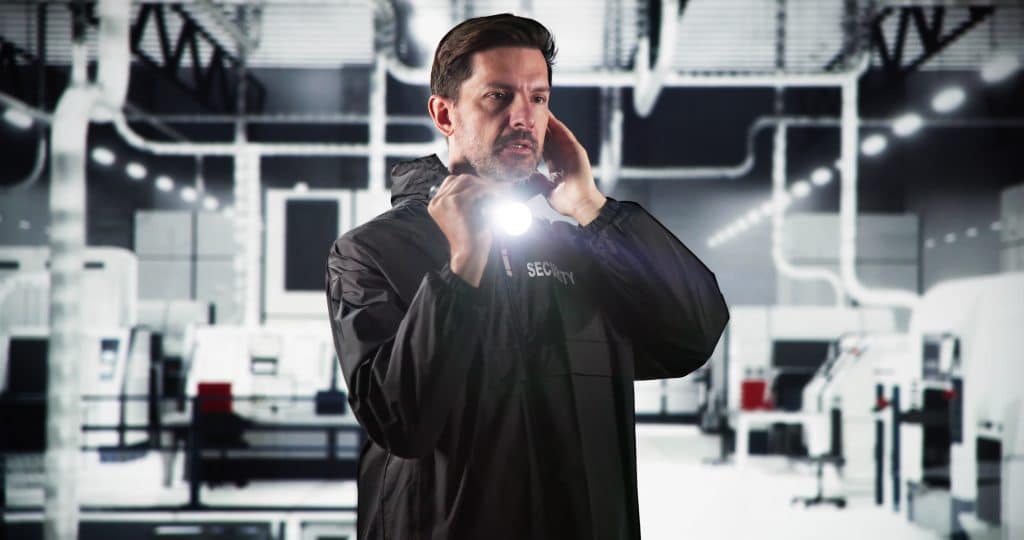 A person wearing a black jacket with "SECURITY" emblazoned on it stands in an industrial setting, representing Security Services Oakland County. They hold a flashlight to their ear, seemingly communicating through it. The backdrop is a modern, well-lit industrial interior.