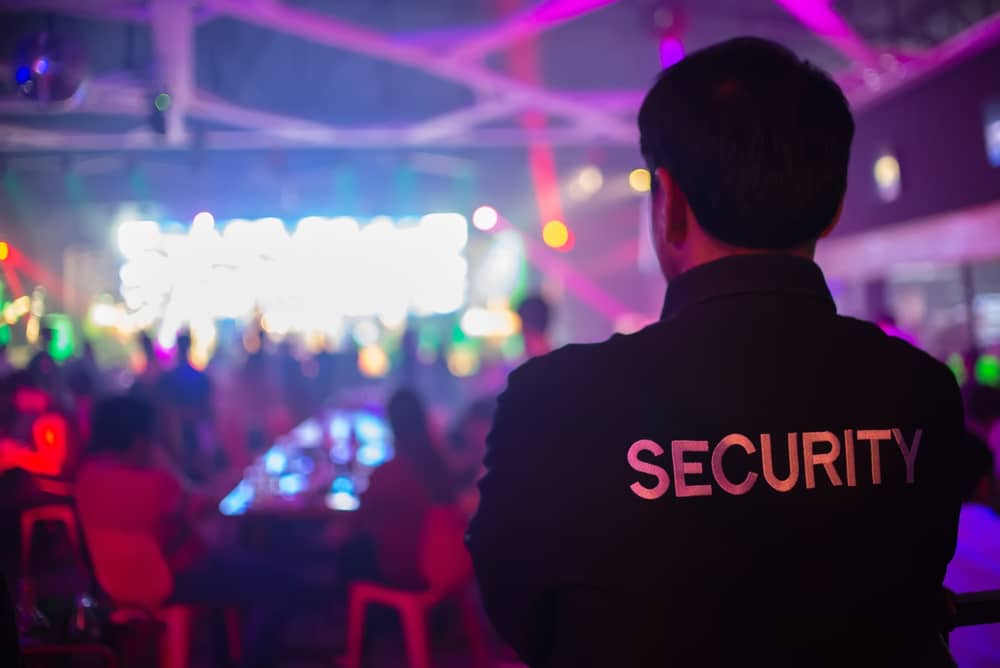 A security guard stands with their back to the camera, wearing a jacket labeled "SECURITY" in a dimly lit nightclub. As vibrant lights dance across the scene, blurred figures mingle—an emblem of the top-tier event security Oakland County trusts for safe and seamless nights.